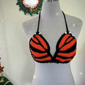 Pode incluir: Top de biqu&iacute;ni de croch&ecirc; feito &agrave; m&atilde;o em listras laranja e pretas, exibido em um manequim. O top halter apresenta acabamento e la&ccedil;os pretos, com pequenos detalhes brancos na parte superior de cada bojo. O design geral assemelha-se a um tigre.