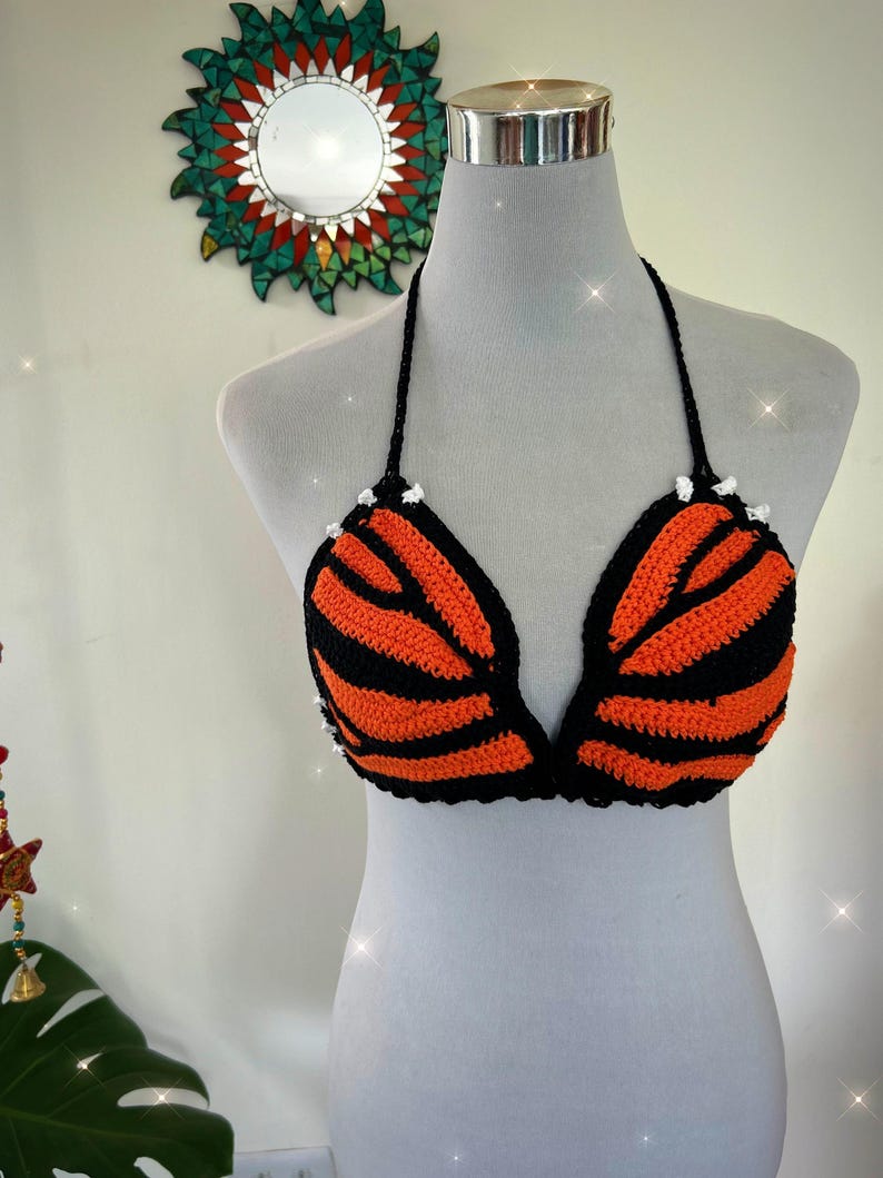 Pode incluir: Top de biqu&iacute;ni de croch&ecirc; feito &agrave; m&atilde;o em listras laranja e pretas, exibido em um manequim. O top halter apresenta acabamento e la&ccedil;os pretos, com pequenos detalhes brancos na parte superior de cada bojo. O design geral assemelha-se a um tigre.