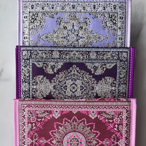 Könnte beinhalten: Drei kleine, rechteckige Gebetsteppiche in Lila- und Rottönen. Jeder Teppich weist ein verziertes, traditionelles Teppichdesign mit goldenen und weißen Akzenten auf. Die Teppiche sind gestapelt und zeigen ihre komplizierten Muster und Texturen.