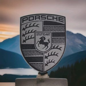 May include: A silver Porsche emblem with the word "PORSCHE" at the top. The emblem features the city name "STUTTGART" and a black horse in the center. The background shows a mountain range.