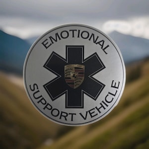 Könnte beinhalten: Ein silberner runder Aufkleber mit der Aufschrift "EMOTIONAL SUPPORT VEHICLE", die ein schwarzes Lebenssternsymbol umgibt. In der Mitte befindet sich ein Porsche-Wappen. Der Hintergrund ist eine verschwommene Außenszene.