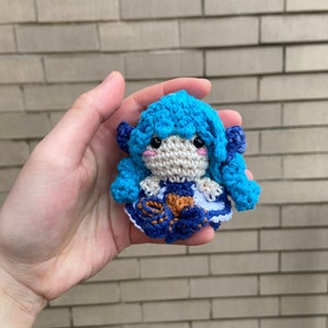 Może przedstawiać: Lalka szydełkowa z niebieskimi włosami, białą sukienką i niebiesko-pomarańczową spódnicą. Lalka trzyma w rękach mały pomarańczowy przedmiot.