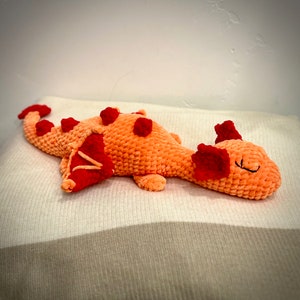 May include: A plush orange dragon toy with red accents, including spikes and wings. The dragon is lying down on a white surface, with a smiling face and black stitched details. The toy is made of a soft, textured material.