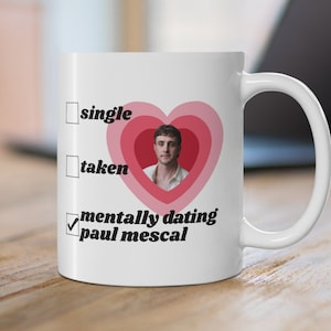Op de afbeelding: Witte keramische mok met een handvat. De mok heeft een hartvormig ontwerp met een foto van Paul Mescal en de tekst "mentally dating paul mescal". De mok heeft de woorden "single" en "taken" met selectievakjes.