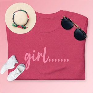 May include: A pink t-shirt with the text "girl......" in pink lettering. The shirt is laid flat on a pink background with a straw hat, sunglasses, and white sneakers.