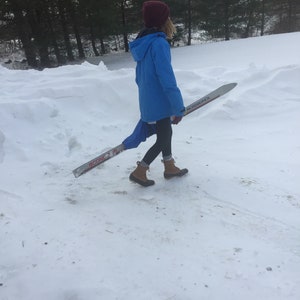 May include: A person in a blue jacket and a red hat walks through a snowy landscape carrying a pair of skis with the text "ROSSIGNOL" and "ICONE 3" printed on them.