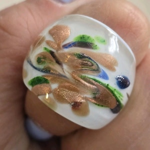 May include: A white glass ring with a square top. The ring features a design of gold, green, and blue swirls and shapes. The ring is a unique piece of jewelry.