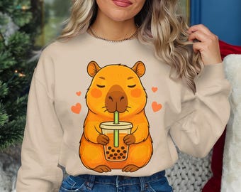 May include: Beige sweatshirt featuring a cartoon capybara drinking boba tea. The capybara is orange with a light brown nose and is surrounded by orange hearts. The boba tea is in a clear cup with a green straw.