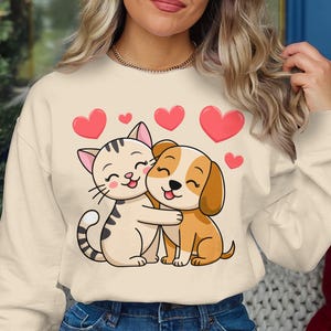 May include: Cream-colored sweatshirt featuring a cartoon cat and dog hugging, surrounded by pink hearts. The cat is light beige with black stripes, and the dog is brown and white. A cheerful design for pet lovers.