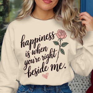 Może przedstawiać: Kremowa bluza z napisem "happiness is when you're right beside me" czarnym pismem. Po prawej stronie tekstu znajduje się różowa róża z zielonymi liśćmi, a pod spodem różowe serce.