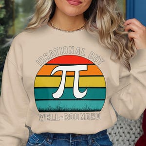 Peut inclure: Sweat-shirt beige avec un motif rétro représentant un coucher de soleil coloré, le symbole pi et le texte "IRRATIONAL BUT WELL-ROUNDED". Le motif est blanc, orange, jaune, vert et bleu.