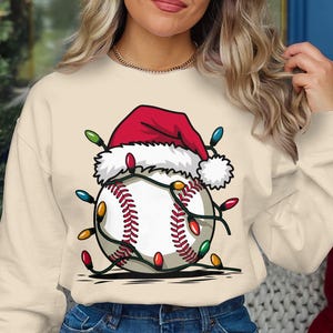 Pode incluir: Camisola creme com um design de basebol com um gorro de Pai Natal e luzes de Natal. O basebol tem costuras vermelhas e é adornado com luzes coloridas. O gorro de Pai Natal é vermelho com uma borda branca.