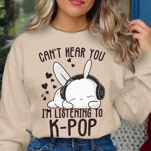 May include: Beige sweatshirt with the text "CAN'T HEAR YOU I'M LISTENING TO K-POP". The graphic features a white bunny wearing headphones and hearts. The sweatshirt is a neutral color.