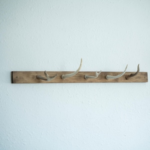 May include: A rustic wooden coat rack with five antler hooks. The rack is made of a weathered, light brown wood and is mounted on a white wall. The antlers are a natural off-white color, adding a touch of nature-inspired decor.