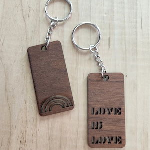 Puede incluir: Dos llaveros de madera con anillas y cadenas plateadas. Un llavero presenta un diseño de arcoíris tallado, y el otro dice "LOVE IS LOVE" en letras recortadas. Los llaveros son de madera marrón oscuro.