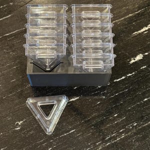 Op de afbeelding: Een set van doorzichtige, driehoekige containers van plastic, gestapeld in twee kolommen op een zwarte basis. Een enkele driehoekige container ligt op het zwarte oppervlak.