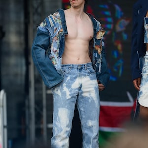 May include: A person wearing a blue denim jacket with patches and light blue jeans with a bleach-splattered design. The person is standing on a stage with a red, green, and black flag in the background.