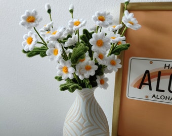 May include: A white ceramic vase with a wavy pattern design holds a bouquet of white daisies with yellow centers. The vase is on a white surface next to a framed picture with the text "Aloha" on it.