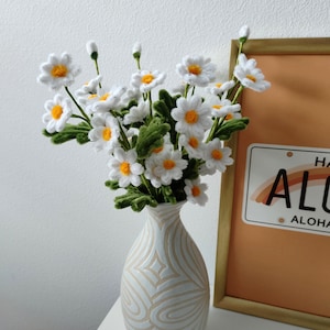 May include: A white ceramic vase with a wavy pattern design holds a bouquet of white daisies with yellow centers. The vase is on a white surface next to a framed picture with the text "Aloha" on it.