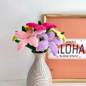 May include: A bouquet of colorful, handmade flowers in a cream-colored vase with a floral design. The flowers are pink, purple, yellow, and fuchsia. A framed print of a Hawaiian license plate is in the background.