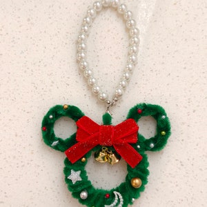 May include: A green Christmas wreath shaped like Mickey Mouse ears with a red velvet bow and gold bells. The wreath is decorated with white pearls, gold beads, and silver stars and moons. A white pearl necklace hangs from the top of the wreath.