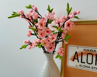 May include: A white ceramic vase with a raised floral design holds a bouquet of pink faux flowers with green stems. The vase is on a white surface in front of a framed print with the text "ALOHA STATE" and a rainbow graphic.