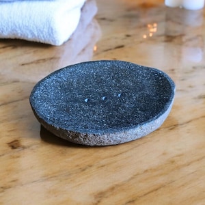 May include: A dark gray stone soap dish with a rough texture and three small holes. The dish is sitting on a wooden surface.
