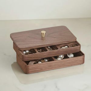 May include: A two-tiered wooden jewellery box with a hinged lid and a small gold-coloured knob. The box is filled with various pieces of jewellery, including pearls and rings. The wood has a warm, natural tone.
