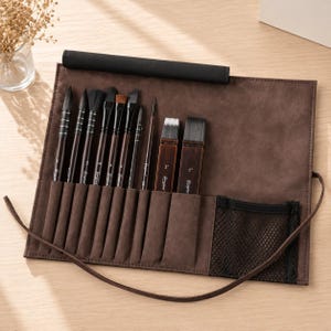May include: A brown fabric roll-up case containing a set of artist paintbrushes with dark wooden handles and black bristles. The case features individual slots for each brush, a black top flap, a black mesh pocket, and a long brown tie for closure.