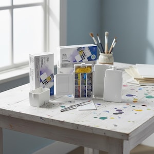 May include: A watercolour painting set with a white plastic case, paint tubes, and a brush. The set includes a white plastic palette, a small white container, and a box with the text "Cotman Watercolour". The items are on a white wooden table.