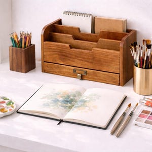 Wooden desktop organiser with three compartments and open drawer on a white table styled with an open sketchbook watercolor paints brushes wooden brush holders and art tools creating a calm artist workspace