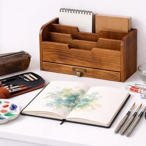 Vintage wooden desk organiser styled with sketchbook watercolor paints brushes and art tools on a white table