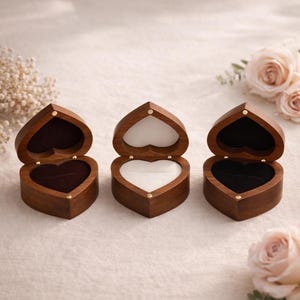 May include: Three heart-shaped wooden ring boxes with hinged lids. The boxes are open, revealing a velvet or felt interior in brown, white, and black. The boxes are made of dark wood and are displayed on a light-coloured surface.