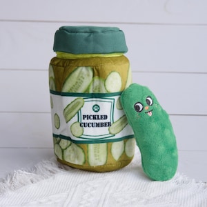 Puede incluir: Un juguete de peluche con forma de tarro de pepinillos encurtidos. El tarro tiene una tapa verde y una etiqueta blanca que dice "Pepinillo encurtido". Hay un juguete de peluche separado con forma de pepinillo verde con una cara sonriente.