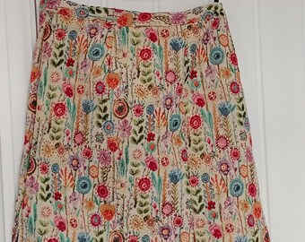 Pode incluir: Uma saia de cor creme com estampa floral que apresenta flores e folhas coloridas. A saia tem uma silhueta reta e uma leve forma de A.