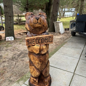 May include: A carved wooden bear sculpture holding a sign that says "WELCOME". The bear is brown and has a friendly expression. The sculpture is standing on a concrete patio.
