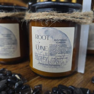 May include: Several glass jars with black lids and labels that read "ROOT LUNE spiritual cleanse." The jars are wrapped with twine and sit on a wooden surface with black stones. The labels include text about essential oils.