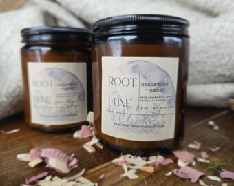May include: Two amber glass candle jars with black lids. The candle on the right has a label that reads "ROOT + LUNE cedarwood + amber hand poured with love in Barrie,  handmade 8 oz - 60+ hour burn time etsy.com/shop/rootandlune".