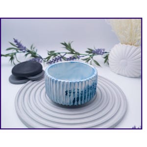 May include: A small, round, blue and white concrete planter with vertical ridges. The planter sits on a gray, circular, ridged tray. Decorative elements include a stack of gray stones, lavender sprigs, and a white seashell-shaped vase.