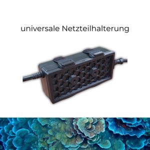 Könnte beinhalten: Ein schwarzer Universal-Netzteilhalter mit Wabenmuster und zwei Klettbändern. Der Text "universale Netzteilhalterung" ist über dem Halter zu sehen. Der Hintergrund zeigt eine Nahaufnahme von blau-grünen Korallen.