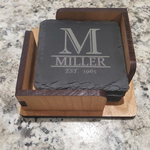 May include: A set of four black slate coasters with a wooden holder. The coasters are engraved with the letter "M", the word "MILLER", and "EST. 1965".