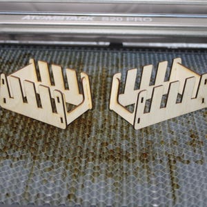 May include: Two identical wooden organizers with a slotted design, likely for holding items. The light-colored wood contrasts with the gray honeycomb surface they rest on. The laser cutting machine is visible in the background.