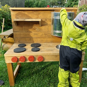 Könnte beinhalten: Eine hölzerne Spielküche mit einem schwarzen Spülbecken, vier schwarzen Kochfeldern und einer Metalldose. Die Küche ist braun lackiert und hat ein kleines Regal. Ein Kind spielt mit der Küche.