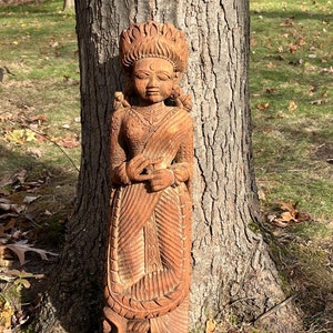 Può includere: Una scultura in legno dettagliata di una figura femminile, scolpita con motivi e disegni intricati. La figura è in piedi, con le mani giunte, e adornata con un copricapo e abiti drappeggiati. La scultura è di colore marrone caldo e si trova vicino a un albero.