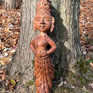 Può includere: Statua in legno intagliato di colore marrone-rossastro raffigurante una figura femminile con corona e dettagli ornati. La statua è posizionata davanti a un albero. Le braccia della figura sono piegate ai gomiti.