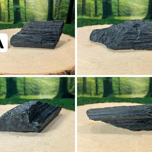 May include: Four raw, black tourmaline stones displayed on a light brown wooden surface. The stones have a rough, textured appearance and vary in shape and size. The background features a blurred forest scene.