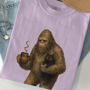 May include: A light purple t-shirt featuring a graphic of a brown, furry Bigfoot creature holding a black cat and a brown mug with the word MEH printed on it. The creature has a neutral facial expression and steam rises from the mug.
