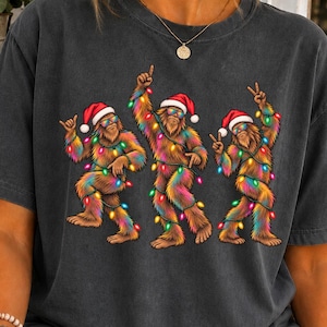 May include: A charcoal gray short-sleeve t-shirt featuring a graphic of three brown Bigfoot creatures wearing Santa hats and sunglasses. Each creature is wrapped in colorful Christmas lights and posed in a dancing position.