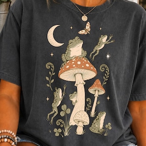 May include: A charcoal gray short-sleeve t-shirt featuring a graphic print of several green frogs, a crescent moon, a butterfly, and various mushrooms. The design includes botanical elements like ferns and clover leaves arranged around the fungi.