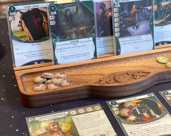 May include: A dark wood card organizer tray with a carved central emblem, holding several game cards upright in a rear slot. The tray contains small circular tokens with graphic symbols, while additional game cards and tokens are arranged on the surface.